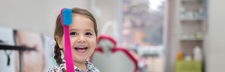 Young girl with braided pigtail smiling and holding a large pink toothbrush with blue bristles