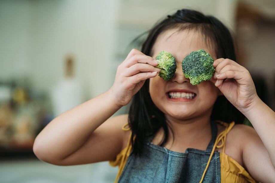 young girl wearing apron putting two broccoli stalks up to her eyes, smiling